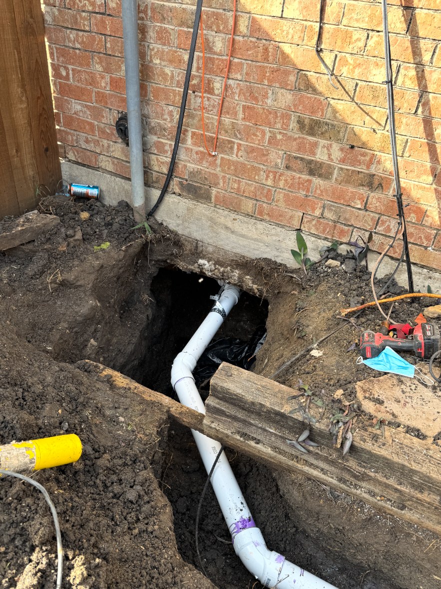 Freshly cut plumbing pipe staged in an excavation trench beside a brick home